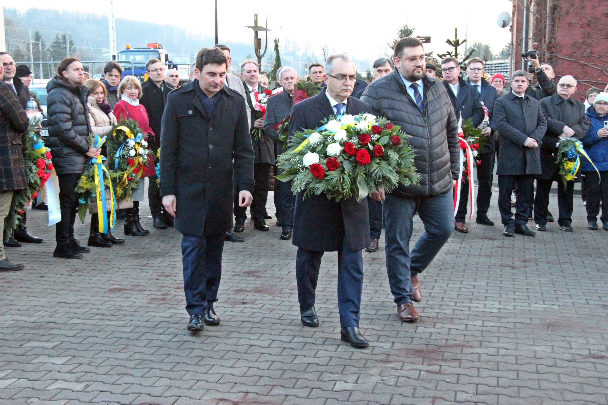 Przedstawiciele urzędu idą z wieńcem do tablicy pamiątkowej